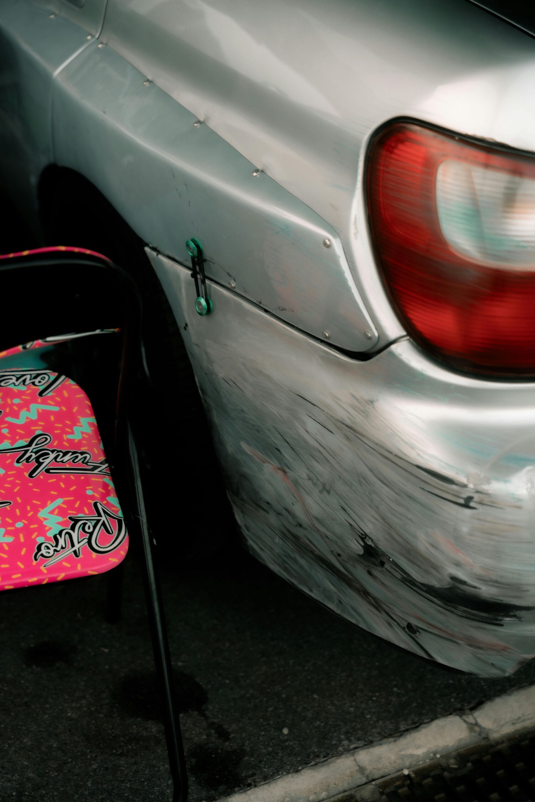 Close-up of a scratched car bumper with a colorful chair beside it outdoors in Cremona, Italy.
